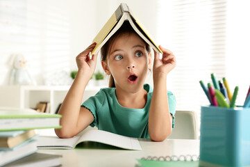 Wall Mural - Emotional little girl with book on her head doing homework at table indoors