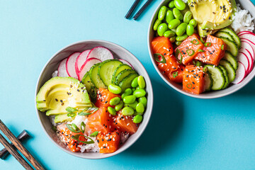 Wall Mural - Poke bowl with salmon, rice, avocado, edamame beans, cucumber and radish in gray bowls, top view. Hawaiian ahi poke bowl, blue background.