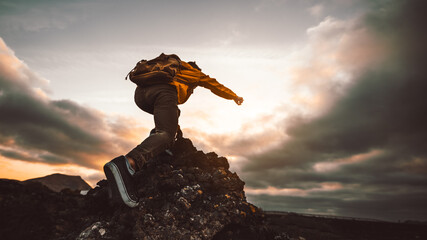 Wall Mural - Hiker standing on the cliff mountain pointing the sky with fingers at sunset. Man on rocky cliff watching down to landscape. Focus on shoe.