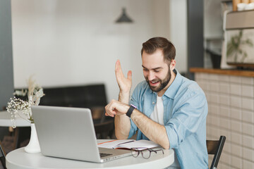 Excited young man sitting alone at table in coffee shop cafe restaurant indoors working or studying on laptop pc computer looking at smart watch on hand. Freelance mobile office business concept.