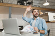 © ViDi Studio - Funny young man sit at table in coffee shop cafe restaurant indoors working studying on laptop pc computer hold papers document waving and greeting with hand. Freelance mobile office business concept.