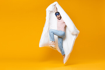 Full length of cheerful laughing young bearded man in pajamas home wear sleep mask jumping hold blanket isolated on bright yellow colour background studio portrait. Relax good mood lifestyle concept.