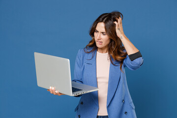 Preoccupied puzzled worried concerned young brunette woman 20s wearing basic jacket standing working on laptop pc computer put hand on head isolated on bright blue colour background studio portrait.