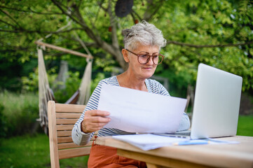 Senior woman with laptop working at the table outdoors in garden, home office concept.