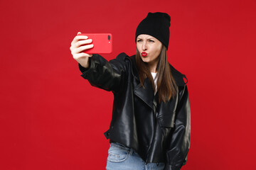 Wall Mural - Pretty cheerful young brunette woman 20s wearing casual black leather jacket white t-shirt hat doing selfie shot on mobile phone making duck face isolated on bright red background studio portrait.