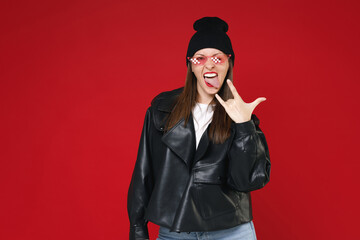 Wall Mural - Funny young brunette woman 20s in black leather jacket white t-shirt hat eyeglasses showing making stick tongue out sign depicting heavy metal rock sign isolated on red background studio portrait.