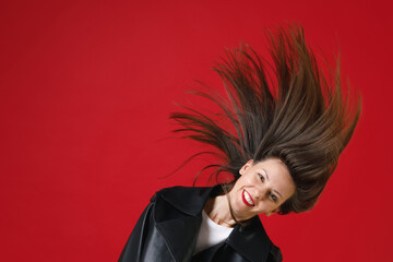 Smiling funny young brunette woman 20s wearing black leather jacket white t-shirt dancing jumping shaking head with fluttering hair looking camera isolated on bright red background studio portrait.