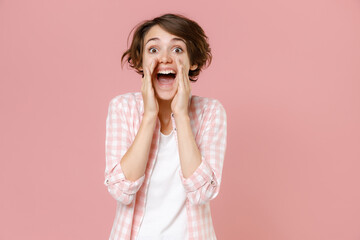 Wall Mural - Excited cheerful young brunette woman 20s wearing casual checkered shirt standing screaming with hands gesture near mouth looking aside isolated on pastel pink colour background, studio portrait.