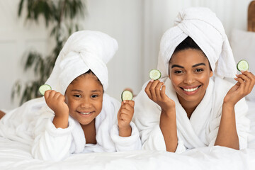 Wall Mural - Beauty Time. African Mom And Daughter In Bathrobes Posing With Cucumber Slices