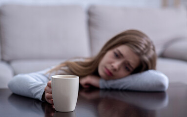 Wall Mural - Sad young woman leaning on table and holding cup with hot drink, suffering from depression. Selective focus. Copy space