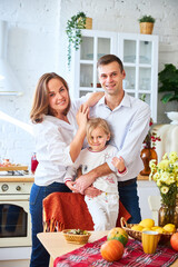 Wall Mural - A happy family of three, mother, father and daughter, stand on a light kitchen, a wooden table with an autumn harvest - pumpkins, apples