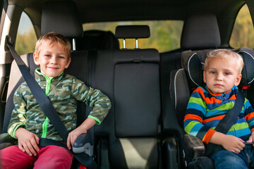 Two little boys sitting on a car seat and a booster seat buckled up in the car.