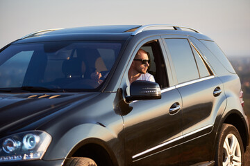 Handsome man is sitting in his black car looking out of the window wearing sunglasses, looking away, outside the city, glimpse of the setting sun, side view