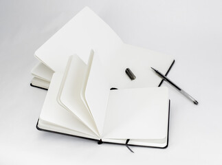 Two black unfold open notebook with blank white pages. Large and small notepads. Dark gel pen with cap. White background