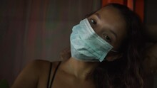 Attractive Long Haired Brunette Young Woman With Brown Eyes Wearing Light Blue Surgical Mask To Prevent Illness Poses For Camera In Dark Room, Close View