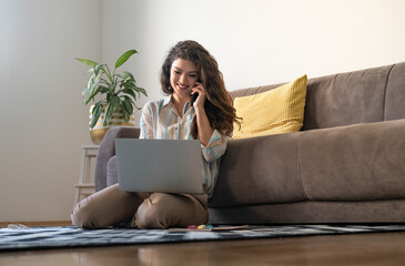 Cheerful woman talking on mobile phone and using laptop computer at home