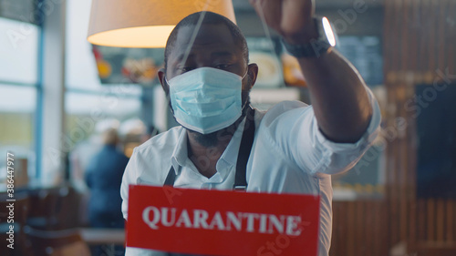 Fotografía Afro-american waiter wearing safety mask hanging quarantine sign in glass door o