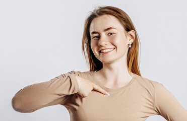 Wall Mural - Beautiful young girl smiling and pointing her her finger at herself on a white isolated background. A woman points to an idea, a place for advertising. Positive brunette in a beige jumper.