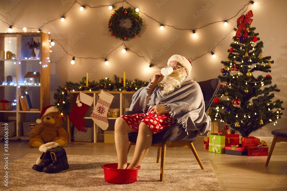 Santa relaxing at home after long day of work, enjoying cup of tea and soaking feet in warm water - obrazy, fototapety, plakaty 