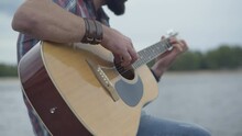 Close-up Of Guitar In Male Caucasian Hands. Unrecognizable Young Man Playing Stringed Musical Instrument With Grey Cloudy Sky And River At The Background.