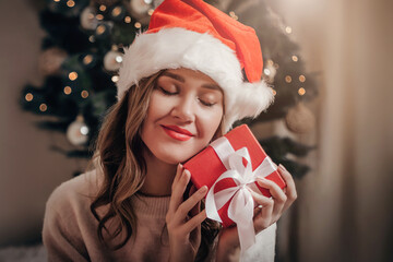 Caucasian girl in a New Year's festive Santa Claus hat holds a gift box near her face and dreams on the background of a Christmas tree and garland. Close up portrait