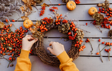 Girl making floral autumn door wreath using colorful rosehip berries, rowan, dry flowers and pumpkins. Fall flower decoration workshop, florist at work.