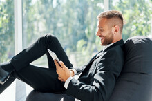 Smiling Successful Businessman In Elegant Suite Using Tablet Sitting On Bean Bag Chair In Creative Office