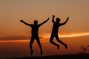 Wall Mural - Silhouette of two Indian friends jumping with arms raised against the sky during the sunset	