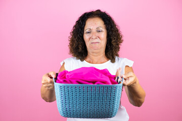 Middle age woman wearing casual white shirt standing over isolated pink background doing housework holding a smelly basket with clothes