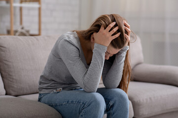 Wall Mural - Unhappy young woman with head in her hands sitting on couch, crying and feeling depressed