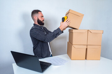 Poster - Storekeeper with a barcode reader in hand. A man accepts goods for storage. Warehouse storage. A bearded man stands at a table with a laptop and cardboard boxes. Working in a warehouse.