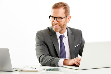 Wall Mural - Studio shot of a smiling businessman wearing suit while sitting at desk and working on laptop. Isolated on white background.