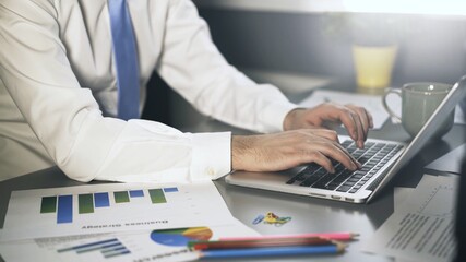 Businessman male hands in white shirt and tie typing in laptop. Man no face texting in laptop on the office table, making project using papers, graphs, pens and pencil