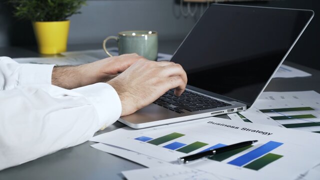 Businessman male hands in white shirt typing in laptop with black screen, graphics and business plan strategy aside. Man hands texting in laptop on the office table, business papers