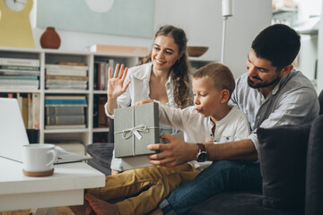 Canvas Print - young happy family with one kid sitting on sofa and opening gifts while having video call on computer