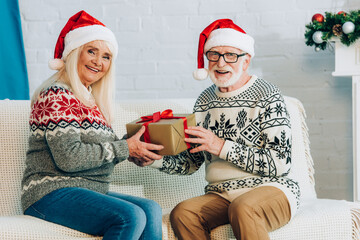 happy senior couple in santa hats looking at camera while sitting on sofa with gift box