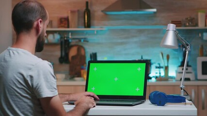 Businessman looking at green screen monitor sitting at home in kitchen. Freelancer watching desktop monitor display with green mockup, chroma key, during night time working overtime.