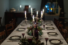 A Dark Romantic Formal Dining Room Table Set With Vintage China Plates, Silver Candelabras With White Lit Candles, A White Tablecloth, Floral Centerpieces, Silverware, And Small Red Wine Glasses. 