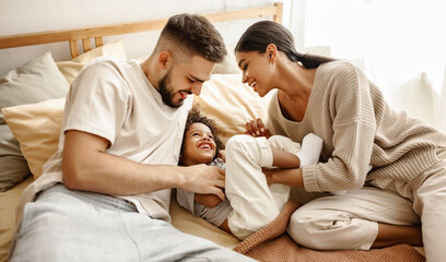 Wall Mural - happy diverse family mom, dad and child  laughing, playing and tickles   in bed   at home.