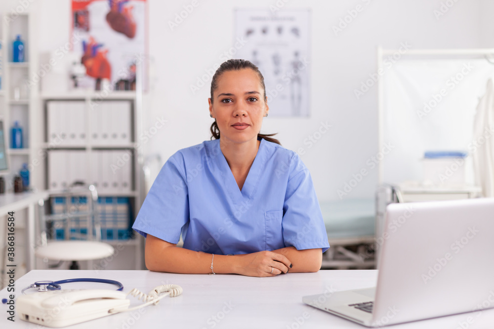Portrait of pretty medical nurse smiling at camera in hospital office wearing blue uniform. Healthcare practitioner sitting at desk using computer in modern clinic looking at monitor, medicine. - obrazy, fototapety, plakaty 