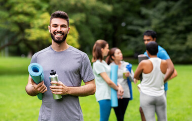 Wall Mural - fitness, sport and healthy lifestyle concept - happy smiling young man with mat and bottle over group of people meeting for yoga class at summer park