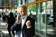 © Carlos David - Portrait of a very excited  senior businessman talking on phone celebrating success holding fist in front of glass building in the city