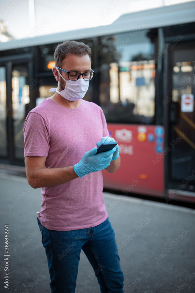 Man with medical protective mask and gloves waiting for a public transportation on a bus-stop and using smartphone. - obrazy, fototapety, plakaty 
