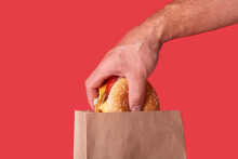 The Guy Pulls Out A Big Juicy Burger From A Craft Brown Ecological Bag. Red Background.