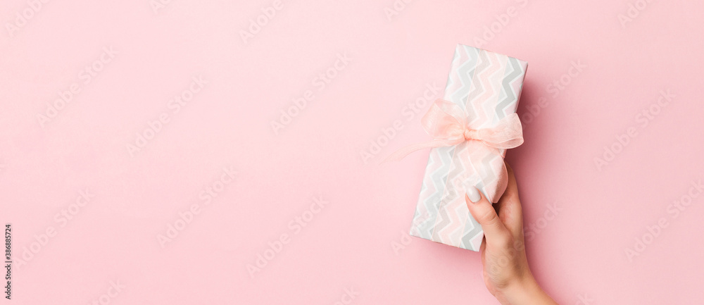 Female's hands holding striped gift box with colored ribbon on living coral background. Christmas concept or other holiday handmade present box, concept top view with copy space - obrazy, fototapety, plakaty 