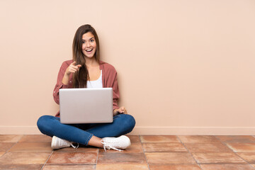 Wall Mural - Teenager student girl sitting on the floor with a laptop surprised and pointing front