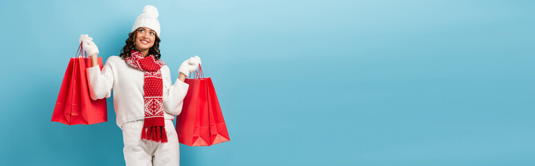 horizontal crop of joyful woman in winter outfit holding red shopping bags on blue