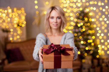 Wall Mural - Christmas and New Year concept - beautiful blonde woman giving Christmas gift in decorated living room with Christmas tree and lights. Focus on gift box