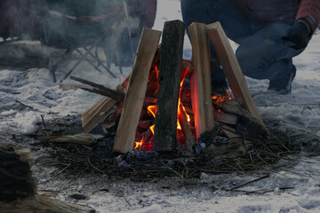 Man is fan the campfire flames in a wild at the dusk, closeup winter camping bonfire at night, front selective focus