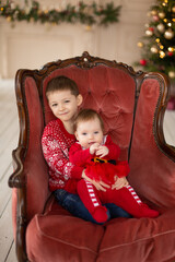 Litttle brother hugs his little sister in red retro chair near Christmas Tree. Enjoying a love hug, people's holidays. Togetherness concept